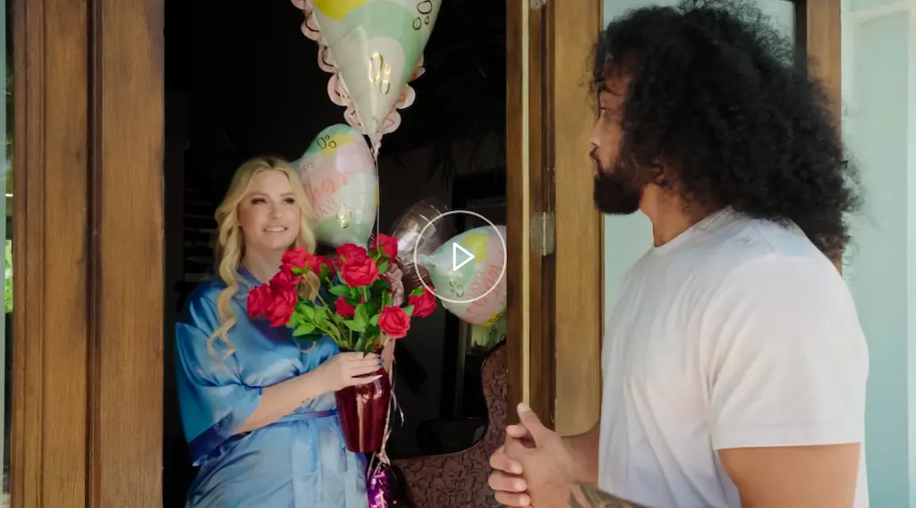 Scena di Lil Humpers 2025 con una sensuale MILF in vestaglia blu che apre la porta con fiori e palloncini, sorpresa dall’arrivo del giovane uomo.